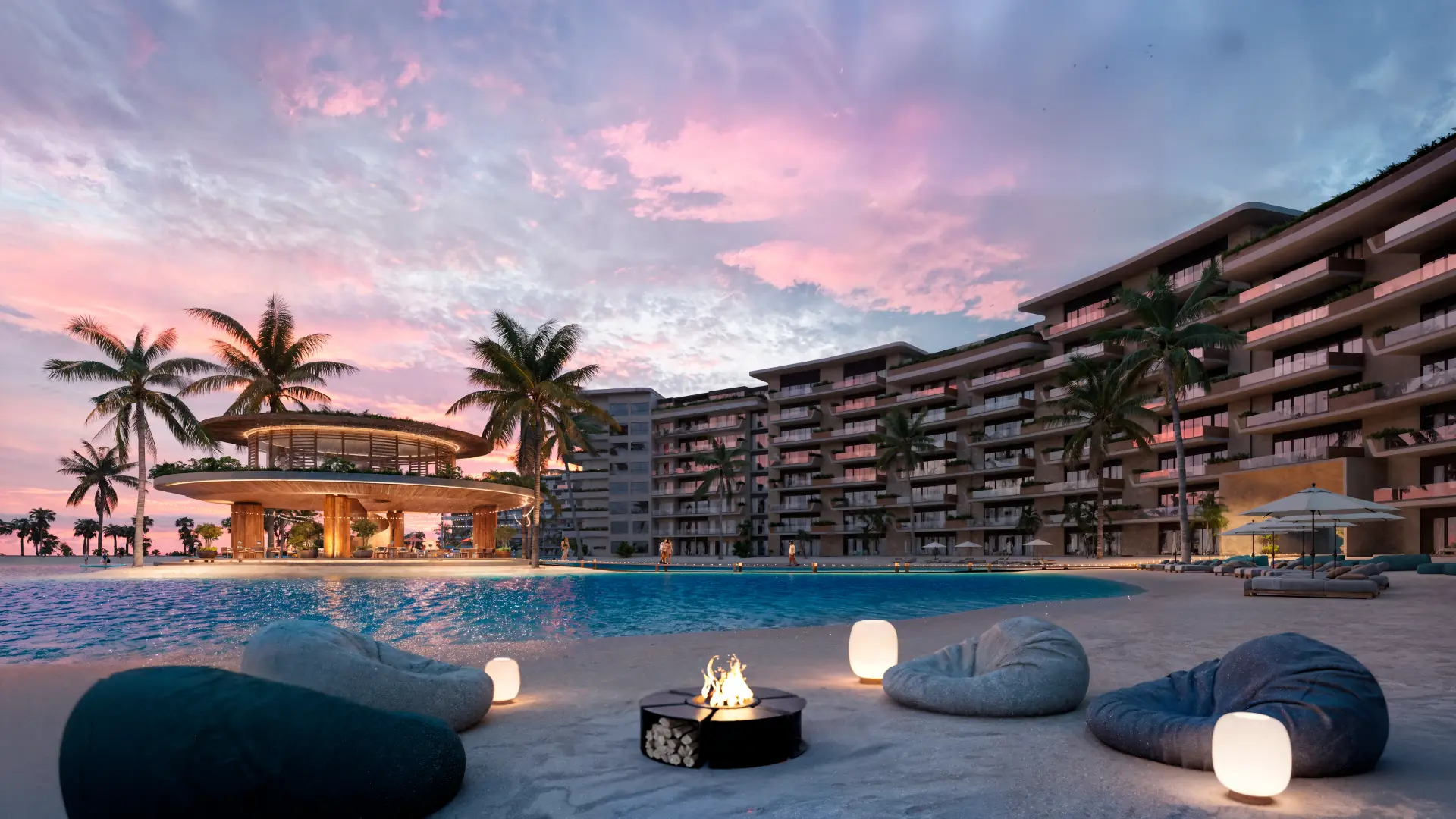 Dusk on the lagoon beach: a pink sky, a fire pit, bean bags and lanterns on the sand, the residences behind.