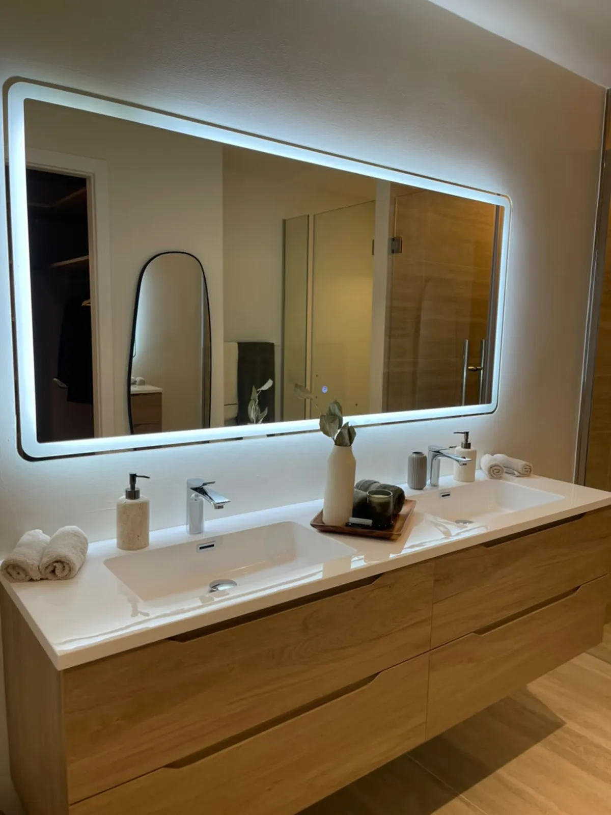 Photographie de la salle de bains de la résidence témoin : miroir éclairé au-dessus d'un plan double vasque en pierre, sur un meuble en bois.